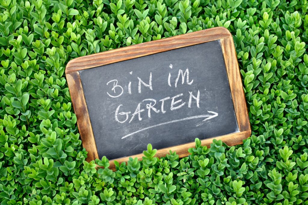 Eine handbeschriftete Tafel mit der Aufschrift 'Bin im Garten', umgeben von dichtem grünen Laub. Perfekt für die Gartendekoration und als Hinweis für Besucher.