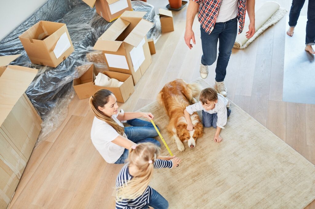 Familie mit Kindern und Hund plant ihren Umzug auf einem Teppich, umgeben von Kartons und Möbeln in einem sonnigen Raum.